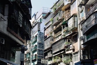 An urban street scene featuring closely-packed residential buildings. Apartments have metal railings and are adorned with potted plants, with air conditioning units attached to many balconies. Sunlight casts shadows, creating a contrast between the bright and dark areas.