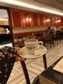 A hand holding a white cup of coffee with intricate designs placed on a matching saucer, alongside a small piece of Turkish delight. The background features a warmly lit cafe with red and gold detailed walls, wooden chairs, and a tiled floor.