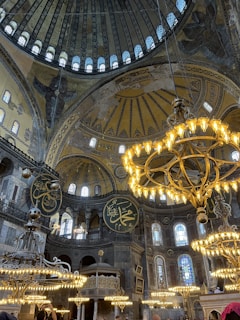 A grand interior with ornate chandeliers hanging from the ceiling. The architecture is elaborate, featuring high domes and intricate artwork, including Arabic calligraphy and decorative patterns on the walls and ceilings. The lighting from the chandeliers adds warmth to the stone structure, highlighting the historic and cultural significance of the space.