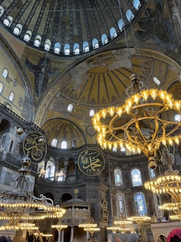 A grand interior with ornate chandeliers hanging from the ceiling. The architecture is elaborate, featuring high domes and intricate artwork, including Arabic calligraphy and decorative patterns on the walls and ceilings. The lighting from the chandeliers adds warmth to the stone structure, highlighting the historic and cultural significance of the space.