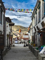 A narrow cobblestone street lined with traditional buildings and colorful prayer flags strung across the alley. In the distance, a large, ornate stupa with orange and white accents stands against a backdrop of rolling mountains and a partly cloudy sky. Local people are casually seated along the street, adding a sense of community and daily life.