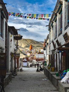 A narrow cobblestone street lined with traditional buildings and colorful prayer flags strung across the alley. In the distance, a large, ornate stupa with orange and white accents stands against a backdrop of rolling mountains and a partly cloudy sky. Local people are casually seated along the street, adding a sense of community and daily life.