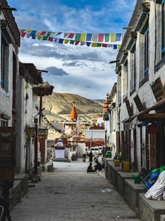 A narrow cobblestone street lined with traditional buildings and colorful prayer flags strung across the alley. In the distance, a large, ornate stupa with orange and white accents stands against a backdrop of rolling mountains and a partly cloudy sky. Local people are casually seated along the street, adding a sense of community and daily life.