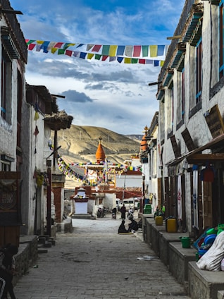 A narrow cobblestone street lined with traditional buildings and colorful prayer flags strung across the alley. In the distance, a large, ornate stupa with orange and white accents stands against a backdrop of rolling mountains and a partly cloudy sky. Local people are casually seated along the street, adding a sense of community and daily life.
