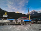 A scenic Himalayan village with traditional stone buildings, adorned with colorful prayer flags and rooftops. Snow-capped mountains rise majestically in the background, partially lit by the golden hues of the setting or rising sun. A stupa or religious structure is present on the left, featuring a golden pinnacle. The scene conveys a peaceful and serene atmosphere.