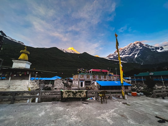 A scenic Himalayan village with traditional stone buildings, adorned with colorful prayer flags and rooftops. Snow-capped mountains rise majestically in the background, partially lit by the golden hues of the setting or rising sun. A stupa or religious structure is present on the left, featuring a golden pinnacle. The scene conveys a peaceful and serene atmosphere.