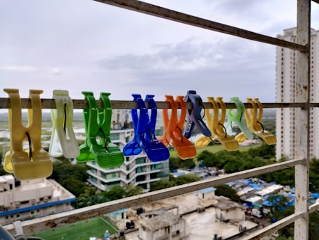 A row of colorful plastic clothespins is clipped onto a metal railing, with a backdrop of a cityscape featuring high-rise buildings and a cloudy sky. The clothespins are various colors including yellow, green, blue, and orange, and are evenly spaced along the railing.