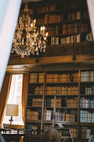 An elegant library with high wooden bookshelves filled with an extensive collection of books. A grand crystal chandelier hangs from the ceiling, adding a touch of luxury. A window with long, golden curtains allows natural light to illuminate the room. A classic lamp sits on a table beside a wooden chair, contributing to the sophisticated ambiance.