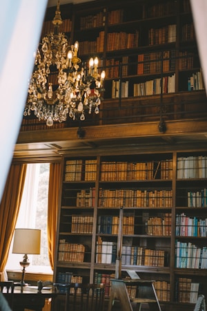 An elegant library with high wooden bookshelves filled with an extensive collection of books. A grand crystal chandelier hangs from the ceiling, adding a touch of luxury. A window with long, golden curtains allows natural light to illuminate the room. A classic lamp sits on a table beside a wooden chair, contributing to the sophisticated ambiance.