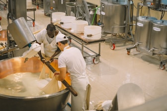 Two people are working together in a cheese-making facility, handling a large copper vat filled with curd. They are wearing white uniforms for hygiene. The machinery and vessels around them suggest an industrial setting. Various metal containers and equipment are visible in the background.