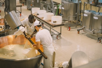 Two people are working together in a cheese-making facility, handling a large copper vat filled with curd. They are wearing white uniforms for hygiene. The machinery and vessels around them suggest an industrial setting. Various metal containers and equipment are visible in the background.