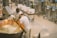 Two people are working together in a cheese-making facility, handling a large copper vat filled with curd. They are wearing white uniforms for hygiene. The machinery and vessels around them suggest an industrial setting. Various metal containers and equipment are visible in the background.