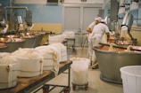 Inside a cheese production facility, two workers wearing white uniforms and caps are tending to large copper vats used for cheese making. Several containers wrapped in cloth are visible on tables and carts, with industrial equipment and pipes lining the walls. The space is clean and organized, reflecting an industrial setting.