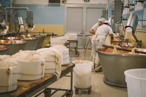 Inside a cheese production facility, two workers wearing white uniforms and caps are tending to large copper vats used for cheese making. Several containers wrapped in cloth are visible on tables and carts, with industrial equipment and pipes lining the walls. The space is clean and organized, reflecting an industrial setting.