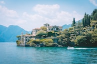 A scenic view of a villa situated on a rocky promontory surrounded by lush greenery and trees. The villa has classical architectural features and overlooks a serene body of water with mountains in the background. A motorboat is gliding across the water in the foreground.