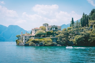 A scenic view of a villa situated on a rocky promontory surrounded by lush greenery and trees. The villa has classical architectural features and overlooks a serene body of water with mountains in the background. A motorboat is gliding across the water in the foreground.