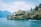 A scenic view of a villa situated on a rocky promontory surrounded by lush greenery and trees. The villa has classical architectural features and overlooks a serene body of water with mountains in the background. A motorboat is gliding across the water in the foreground.