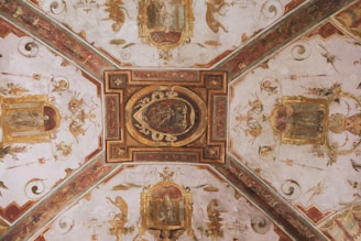 An ornate ceiling mural featuring intricate paintings and decorative motifs with gold embellishments. The design includes symmetrical patterns, mythical figures, and floral elements, all within an architectural framework.