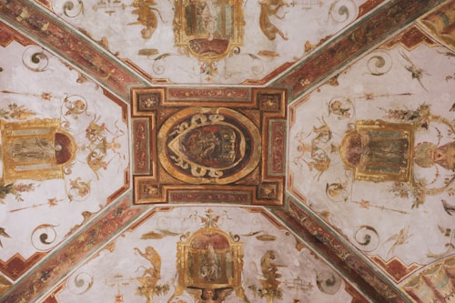 An ornate ceiling mural featuring intricate paintings and decorative motifs with gold embellishments. The design includes symmetrical patterns, mythical figures, and floral elements, all within an architectural framework.