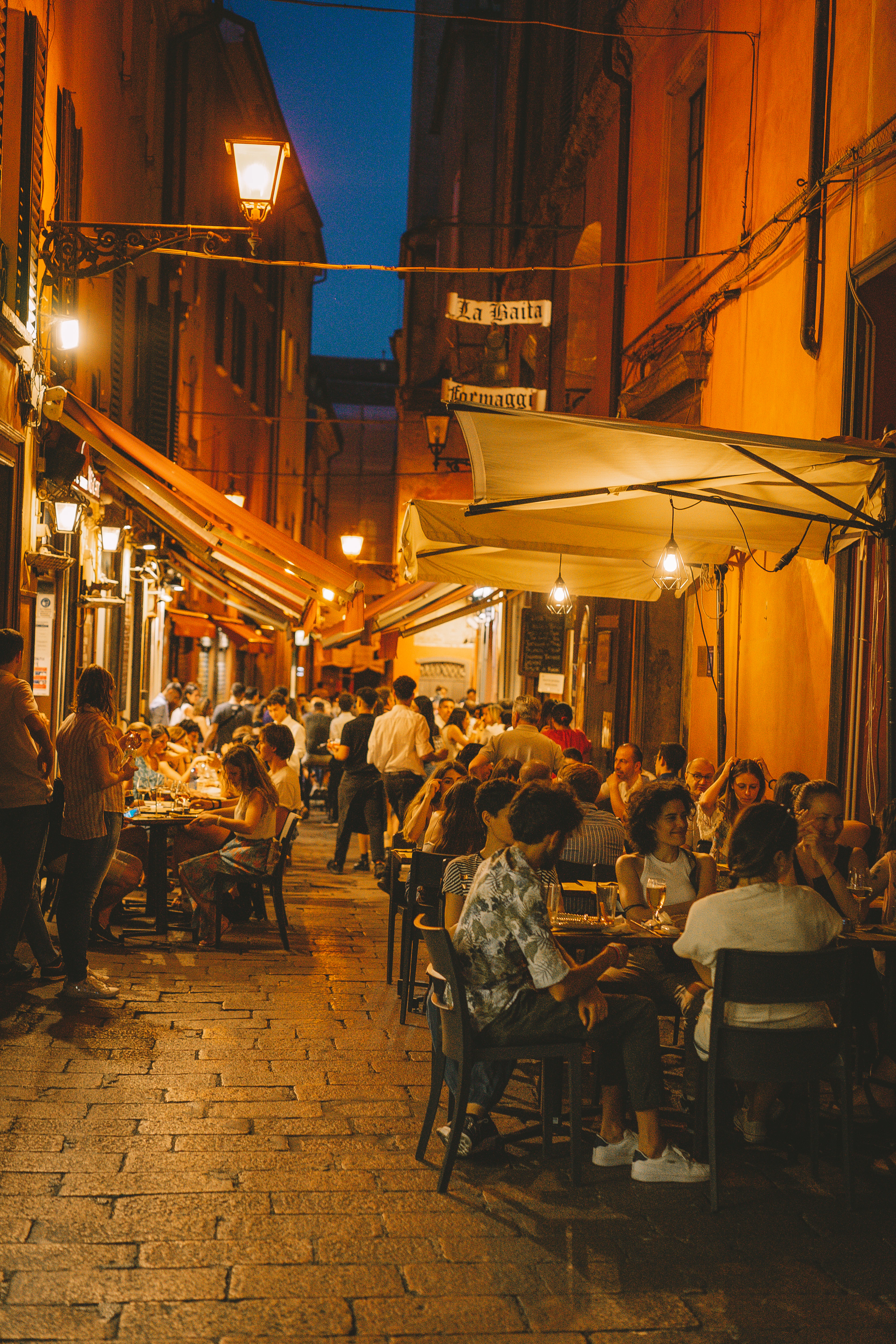 A narrow Bologna alley glowing at night, lantern-lit outdoor tables crowded with diners under striped awnings.
