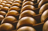Rows of large, round cheese wheels are stacked on wooden shelves, creating a repetitive geometric pattern. The surface of the cheese has a warm, golden-brown hue and is marked with engraved text.