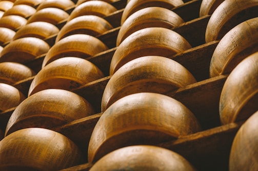Rows of large, round cheese wheels are stacked on wooden shelves, creating a repetitive geometric pattern. The surface of the cheese has a warm, golden-brown hue and is marked with engraved text.