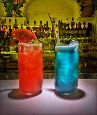 Two vibrant cocktails are placed on a bar counter. One cocktail is red with a slice of dried orange and a sprig of mint as garnish while the other is blue with foam on top and an ornate straw. Behind them, there are numerous bottles of liquor on shelves, creating a colorful background.
