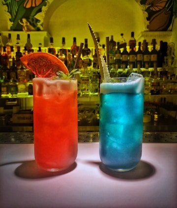Two vibrant cocktails are placed on a bar counter. One cocktail is red with a slice of dried orange and a sprig of mint as garnish while the other is blue with foam on top and an ornate straw. Behind them, there are numerous bottles of liquor on shelves, creating a colorful background.