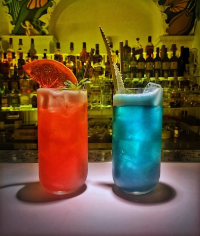 Two vibrant cocktails are placed on a bar counter. One cocktail is red with a slice of dried orange and a sprig of mint as garnish while the other is blue with foam on top and an ornate straw. Behind them, there are numerous bottles of liquor on shelves, creating a colorful background.