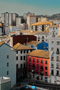 A densely packed urban area with a mix of residential buildings featuring colorful facades, including bright red and blue walls with yellowish roofs. In the background, there are taller, neutral-colored high-rise apartment buildings. The scene is set against a backdrop of hills and partially cloudy sky.