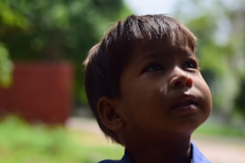 A young child with a contemplative expression looks upward into natural light. The background is blurred with hints of green foliage, suggesting an outdoor setting. Shadows play across the child's face, emphasizing the facial features.