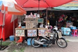 A small coffee stall is set up on the sidewalk, featuring a rustic wooden counter with signs advertising Arabica coffee for sale. Various coffee-related products and a menu are displayed. In front of the stall, a motorbike is parked with a sign saying 'Coffee My Friend.' The background shows a variety of colorful buckets and household items hanging from a shop.