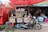 A small coffee stall is set up on the sidewalk, featuring a rustic wooden counter with signs advertising Arabica coffee for sale. Various coffee-related products and a menu are displayed. In front of the stall, a motorbike is parked with a sign saying 'Coffee My Friend.' The background shows a variety of colorful buckets and household items hanging from a shop.