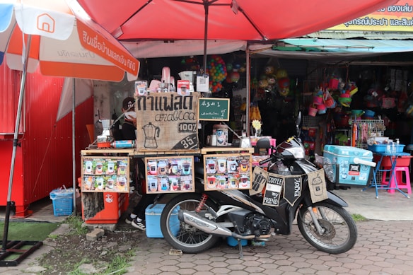 A small coffee stall is set up on the sidewalk, featuring a rustic wooden counter with signs advertising Arabica coffee for sale. Various coffee-related products and a menu are displayed. In front of the stall, a motorbike is parked with a sign saying 'Coffee My Friend.' The background shows a variety of colorful buckets and household items hanging from a shop.