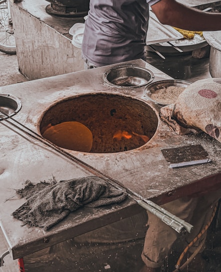 A person is seen working at a traditional clay oven (tandoor), likely baking flatbread. Various kitchen items are scattered around, including metal utensils and containers with ingredients. A stack of plates and a cloth are also visible. The setting appears rustic and busy, indicating an active cooking environment.