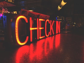 A neon sign displaying the word 'Checkin' in bright red letters on a bar counter. The surrounding environment is dimly lit, with reflections of the sign visible on the surface of the counter. Various bar elements and bottles can be seen faintly in the background.
