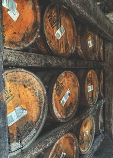 Several wooden barrels are stacked on wooden shelves. The barrels appear aged, with visible labels and marks on their surfaces. The setting suggests a rustic storage area, possibly for aging spirits like whiskey or wine.