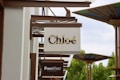 A retail storefront with the brand sign 'Chlo&eacute;' prominently displayed, featuring sleek, golden lettering on a white background. The architectural elements include brown metal frameworks and modern design. Another sign for 'Paw Paws' is visible, suggesting a shopping area. The background shows trees and sky, indicating an outdoor scene.