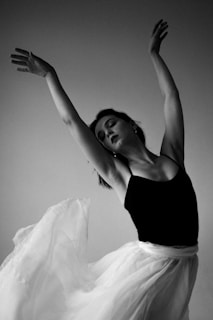 A woman in a graceful dance pose, wearing a black top and a flowing white skirt. Her arms are stretched upwards, and her expression conveys a sense of serenity and focus. The monochrome setting adds to the elegance and timelessness of the scene.