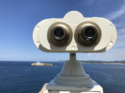A pair of binoculars mounted on a stand overlooks a vast body of water, likely an ocean or sea, with a small island and a lighthouse visible in the distance. The sky is mostly clear, with a few scattered clouds, and the horizon stretches across the background.