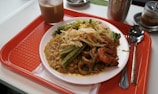 A meal served on a white plate includes a variety of foods such as curry with chickpeas, shrimp, okra, and a mixture of thin noodles and vegetables. The dish is placed on a red cafeteria tray alongside a spoon, fork, and a beverage in a glass. Additional condiments in small jars are visible in the background.