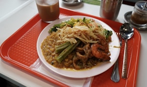 A meal served on a white plate includes a variety of foods such as curry with chickpeas, shrimp, okra, and a mixture of thin noodles and vegetables. The dish is placed on a red cafeteria tray alongside a spoon, fork, and a beverage in a glass. Additional condiments in small jars are visible in the background.