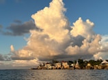 Massive, billowing clouds hover over a coastal village, with a mixture of old and modern buildings nestled among lush greenery. The sunlight casts warm tones on the clouds and the village, while the deep blue sea calmly stretches in the foreground.