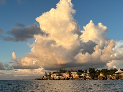 Massive, billowing clouds hover over a coastal village, with a mixture of old and modern buildings nestled among lush greenery. The sunlight casts warm tones on the clouds and the village, while the deep blue sea calmly stretches in the foreground.