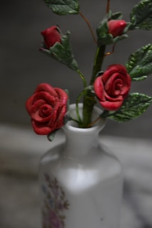 A white vase decorated with floral designs holds several red artificial roses. The roses have green leaves and stems, and the background is blurred with shades of gray.