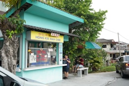A small street food stall with a sign reading 'Hong Kee Mah Chee' is located next to a tree. The stall has a light blue exterior with a yellow sign and clear glass windows displaying various items. Two people are sitting on a bench under an umbrella near the stall, seemingly enjoying their food. Nearby, a car is parked on the road, and residential houses can be seen in the background.