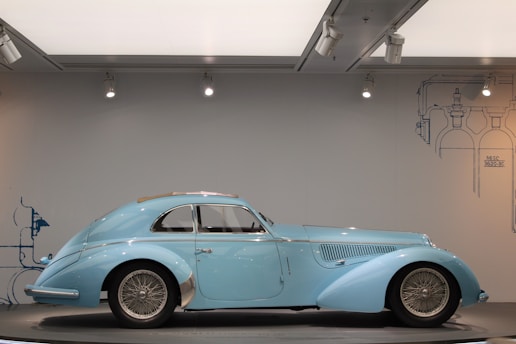 A vintage blue classic car with sleek, aerodynamic lines and wire-spoked wheels is displayed in a well-lit showroom. The car exudes elegance and is positioned against a subtle backdrop with industrial-style blueprints.