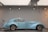 A vintage blue classic car with sleek, aerodynamic lines and wire-spoked wheels is displayed in a well-lit showroom. The car exudes elegance and is positioned against a subtle backdrop with industrial-style blueprints.