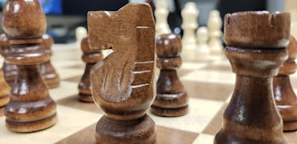A close-up view of a wooden chessboard with several chess pieces, including a prominent knight in the foreground. The pieces are detailed and polished, set on a checkered board with alternating light and dark squares. The background includes blurred elements, suggesting the focus is on the chess pieces.