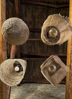 Multiple rustic light fixtures hang from a wooden ceiling, each made from natural materials like twine and burlap. The fixtures feature exposed bulbs, adding to the organic and handmade aesthetic, set against a backdrop of dark wooden planks.